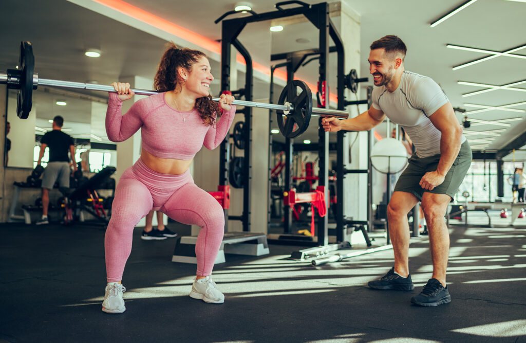 A strong muscular sportswoman is squatting in a gym with a barbell on her shoulders and smiling at her trainer who is helping her. A positive atmosphere in s gym with a personal trainer.